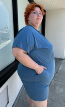 Load image into Gallery viewer, Side view of a size L Universal Standard dusty blue stretchy romper with pockets styled with white platform sandals on a size 22/24 model. The photo was taken outside in natural lighting.
