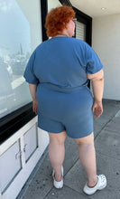 Load image into Gallery viewer, Full-body back view of a size L Universal Standard dusty blue stretchy romper with pockets styled with white platform sandals on a size 22/24 model. The photo was taken outside in natural lighting.
