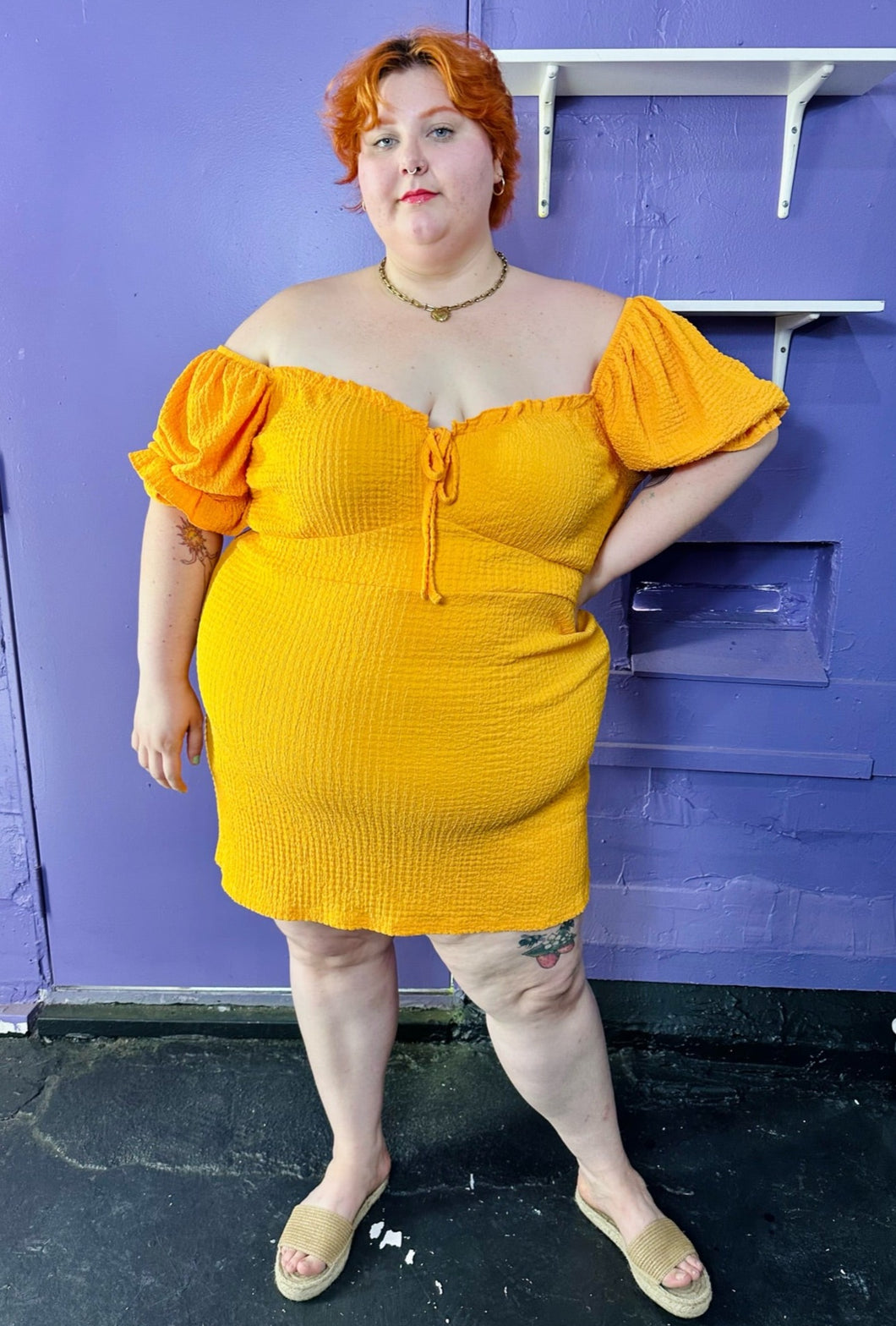 Full-body front view of a size 24 Wednesday's Girl golden yellow textured off-shoulder puff sleeve mini dress with sweetheart neckline styled with tan sandals on a size 22/24 model. The photo was taken inside in fluorescent and natural lighting.