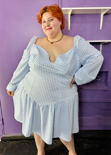 Front view of a size 24 ASOS baby blue and white gingham off-shoulder pointed waistline mini dress on a size 22/24 model. The photo was taken inside in fluorescent and natural lighting.
