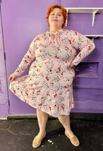 Load image into Gallery viewer, Full-body front view of a size 18 Eloquii baby pink long sleeve midi dress with red, yellow, and blue mini floral print long sleeve midi dress with tie neck detail on a size 22/24 model. The photo was taken inside in flourescent and natural lighting.
