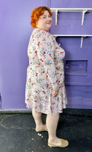 Load image into Gallery viewer, Full-body side view of a size 18 Eloquii baby pink long sleeve midi dress with red, yellow, and blue mini floral print long sleeve midi dress with tie neck detail on a size 22/24 model. The photo was taken inside in flourescent and natural lighting.
