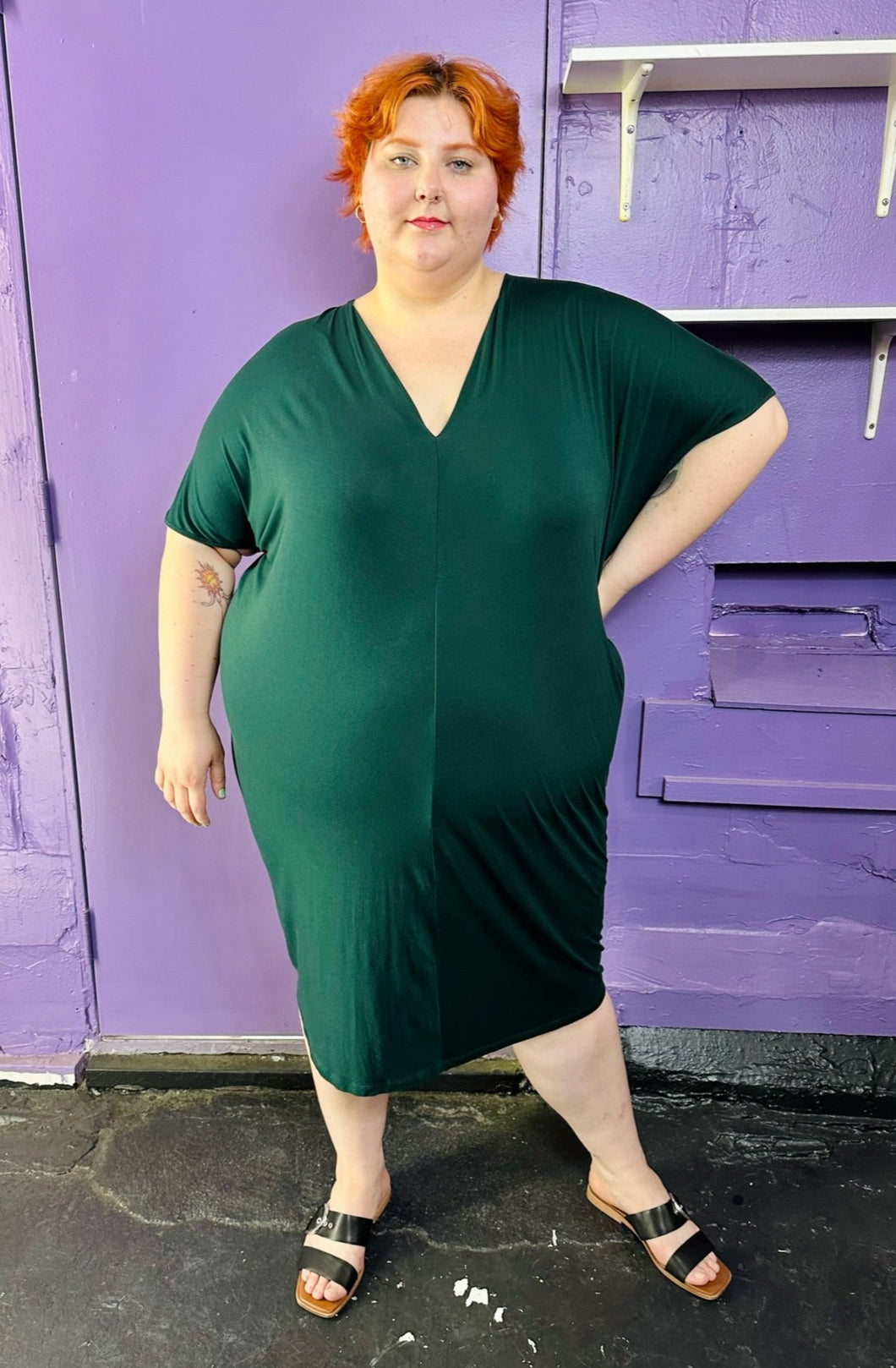 Full-body front view of a size L Universal Standard emerald green v-neck caftan midi dress styled with black and brown sandals on a size 22/24 model. The photo was taken inside under flourescent and natural lighting.