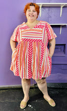 Load image into Gallery viewer, Full-body front view of a size 3X Old Navy white, pink, and orange geometric print a-line mini dress styled with tan sandals on a size 22/24 model. The photo was taken inside in flourescent and natural lighting.
