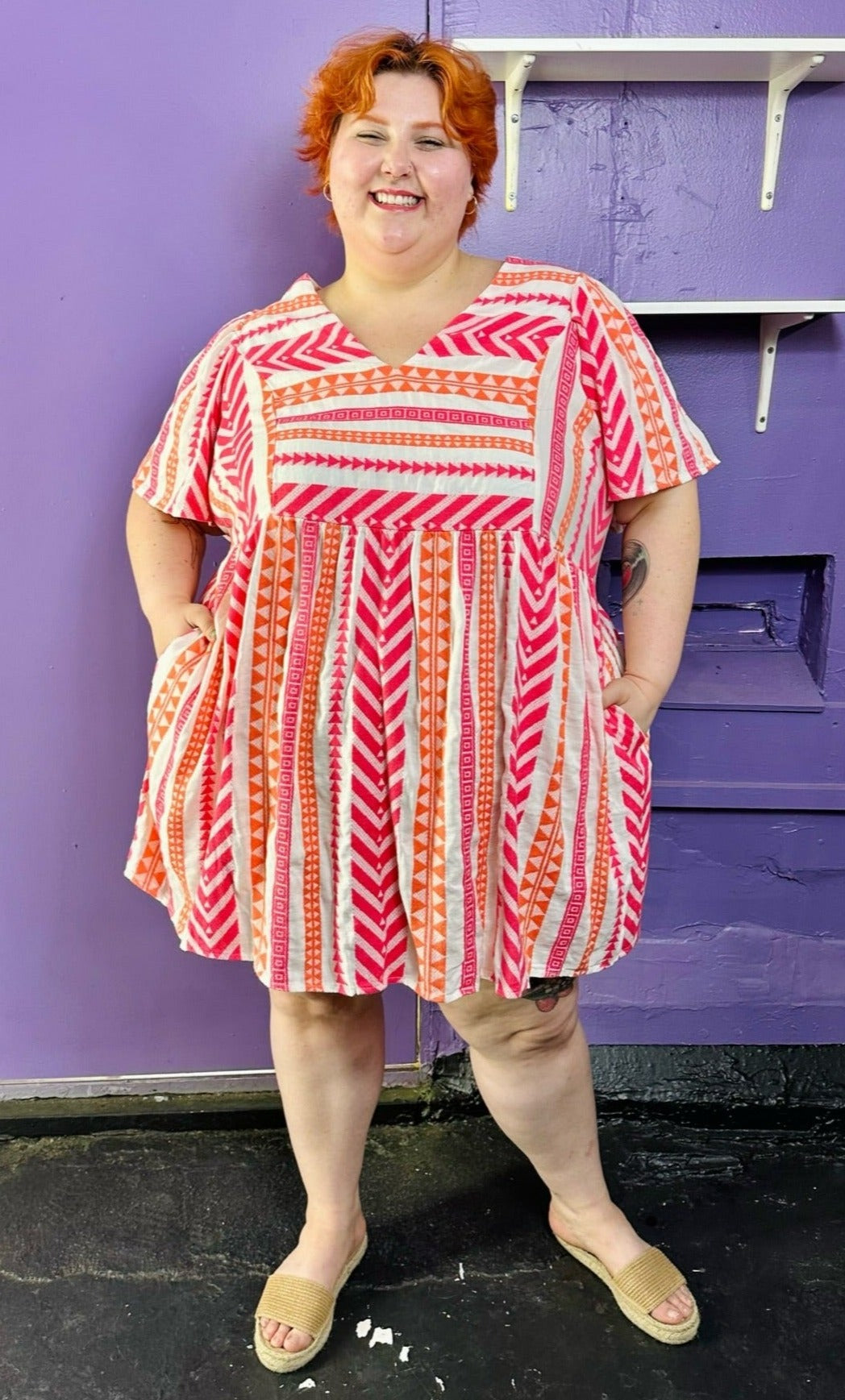 Full-body front view of a size 3X Old Navy white, pink, and orange geometric print a-line mini dress styled with tan sandals on a size 22/24 model. The photo was taken inside in flourescent and natural lighting.