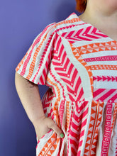 Load image into Gallery viewer, Close up view of the embroidered geometric pattern of a size 3X Old Navy white, pink, and orange geometric print a-line mini dress styled with tan sandals on a size 22/24 model. The photo was taken inside in flourescent and natural lighting.
