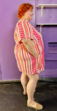 Load image into Gallery viewer, Full-body side view of a size 3X Old Navy white, pink, and orange geometric print a-line mini dress styled with tan sandals on a size 22/24 model. The photo was taken inside in flourescent and natural lighting.
