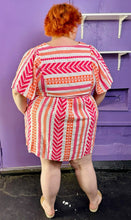 Load image into Gallery viewer, Full-body back view of a size 3X Old Navy white, pink, and orange geometric print a-line mini dress styled with tan sandals on a size 22/24 model. The photo was taken inside in flourescent and natural lighting.
