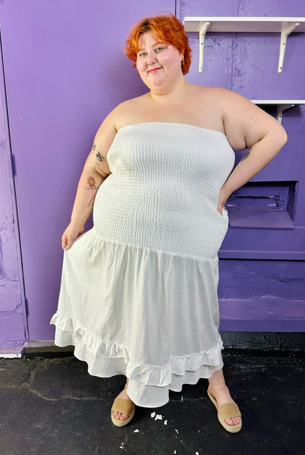 Full-body front view of a size 26 Eloquii white strapless all-over smocked column dress with tiered ruffle hem styled with tan slides on a size 22/24 model. The photo was taken inside under fluorescent and natural lighting.