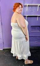 Load image into Gallery viewer, Full-body side view of a size 26 Eloquii white strapless all-over smocked column dress with tiered ruffle hem styled with tan slides on a size 22/24 model. The photo was taken inside under fluorescent and natural lighting.
