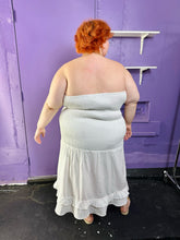 Load image into Gallery viewer, Full-body back view of a size 26 Eloquii white strapless all-over smocked column dress with tiered ruffle hem styled with tan slides on a size 22/24 model. The photo was taken inside under fluorescent and natural lighting.
