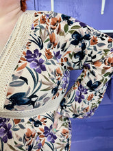 Load image into Gallery viewer, Close up view of the crepe material and gorgeous floral pattern of a size 3 Torrid light tan, orange, purple, and black floral crochet embroidered detail 70s inspired long sleeve midi dress on a size 22/24 model. The photo was taken inside in flourescent and natural lighting.
