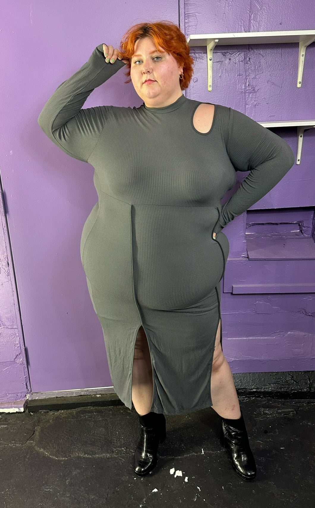Full-body front view of a size 3X Finesse dark gray ribbed long sleeve midi dress with a shoulder cut out, two front slits, and contrast stitching details styled with patent leather black boots on a size 22/24 model. The photo was taken inside in flourescent and natural lighting.