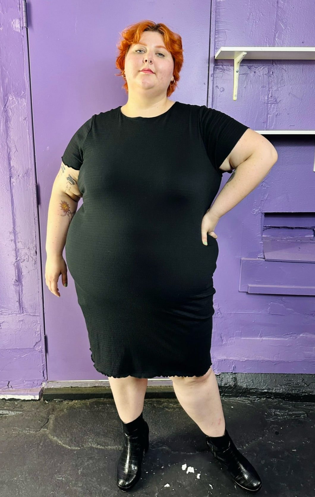 Full-body front view of a size 3 Torrid black sleeveless textured shift midi dress styled with black patent leather boots on a size 22/24 model. The photo was taken inside under flourescent and natural lighting.