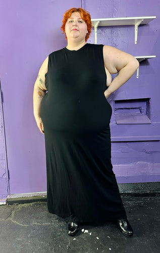 Full-body front view of a size 22 ASOS black sleeveless column maxi dress styled with black boots on a size 22/24 model. The photo was taken inside under flourescent and natural lighting.