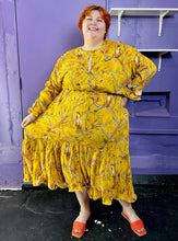 Load image into Gallery viewer, Full-body front view of a size 4X H&amp;M x Johanna Ortiz golden yellow, white, and brown mixed pattern of birds in trees maxi dress with keyhole neckline and back, ruffle hem, and ruffles cuffs styled with orange sandals on a size 22/24 model. The photo was taken inside under flourescent and natural lighting.

