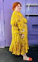 Load image into Gallery viewer, Full-body side view of a size 4X H&amp;M x Johanna Ortiz golden yellow, white, and brown mixed pattern of birds in trees maxi dress with keyhole neckline and back, ruffle hem, and ruffles cuffs styled with orange sandals on a size 22/24 model. The photo was taken inside under flourescent and natural lighting.
