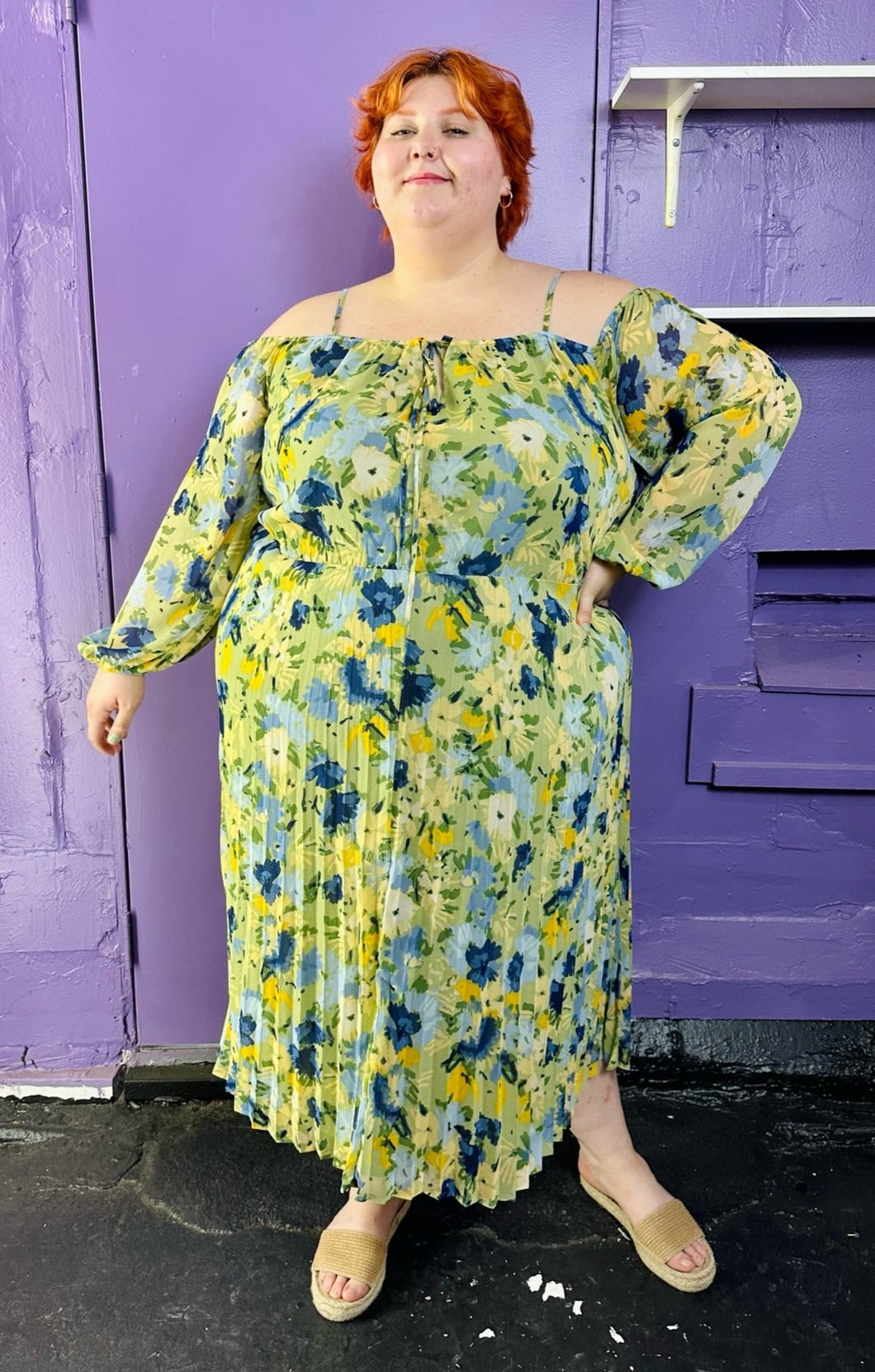 Full-body front view of a size 24 Eloquii light green, yellow, and dark and light blue elegant floral pattern off-the-shoulder / cold-shoulder maxi dress with pleated skirt detail styled with tan slides on a size 22/24 model. The photo was taken inside under fluorescent and natural lighting. 