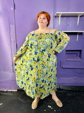 Load image into Gallery viewer, Additional full-body front view of a size 24 Eloquii light green, yellow, and dark and light blue elegant floral pattern off-the-shoulder / cold-shoulder maxi dress with pleated skirt detail styled with tan slides on a size 22/24 model. The photo was taken inside under fluorescent and natural lighting. 
