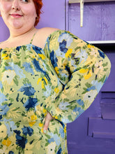 Load image into Gallery viewer, Close up view of the off-the-shoulder / cold-shoulder silhouette and painterly floral pattern of a size 24 Eloquii light green, yellow, and dark and light blue elegant floral pattern off-the-shoulder / cold-shoulder maxi dress with pleated skirt detail on a size 22/24 model. The photo was taken inside under fluorescent and natural lighting. 
