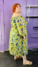 Load image into Gallery viewer, Full-body side view of a size 24 Eloquii light green, yellow, and dark and light blue elegant floral pattern off-the-shoulder / cold-shoulder maxi dress with pleated skirt detail styled with tan slides on a size 22/24 model. The photo was taken inside under fluorescent and natural lighting. 
