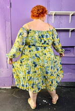 Load image into Gallery viewer, Full-body back view of a size 24 Eloquii light green, yellow, and dark and light blue elegant floral pattern off-the-shoulder / cold-shoulder maxi dress with pleated skirt detail styled with tan slides on a size 22/24 model. The photo was taken inside under fluorescent and natural lighting. 
