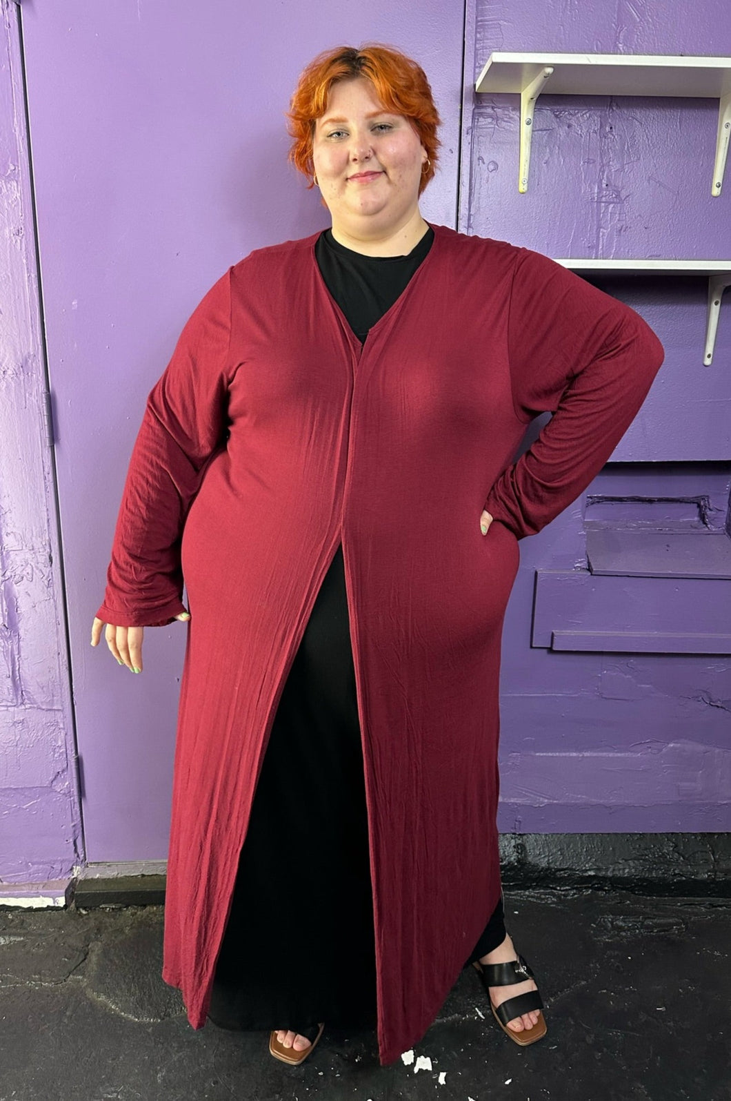 Full-body front view of a size XXXL Zelie for She maroon long sleeve longline duster styled closed over a black maxi dress on a size 22/24 model. The photo was taken inside in fluorescent and natural lighting.