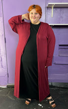 Load image into Gallery viewer, Full-body front view of a size XXXL Zelie for She maroon long sleeve longline duster styled open over a black maxi dress on a size 22/24 model. The photo was taken inside in fluorescent and natural lighting.
