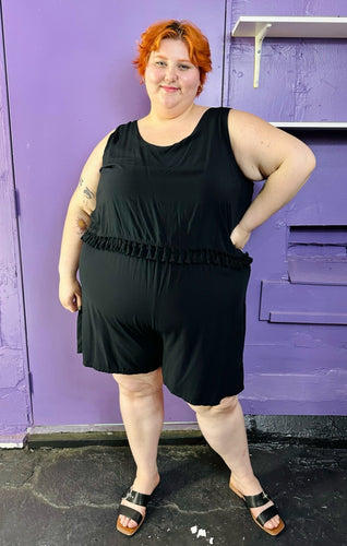 Full-body front view of a size 28 Lane Bryant black tank romper with fringe waist detail styled with black and brown sandals on a size 22/24 model. The photo was taken inside under fluorescent and natural lighting.