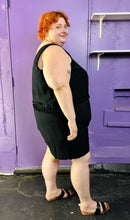 Load image into Gallery viewer, Full-body side view of a size 28 Lane Bryant black tank romper with fringe waist detail styled with black and brown sandals on a size 22/24 model. The photo was taken inside under fluorescent and natural lighting.
