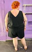 Load image into Gallery viewer, Full-body back view of a size 28 Lane Bryant black tank romper with fringe waist detail styled with black and brown sandals on a size 22/24 model. The photo was taken inside under fluorescent and natural lighting.
