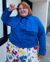 Load image into Gallery viewer, Additional front view of a size 3X Target x Black History Month cobalt blue wind breaker with large silver snap-closure buttons styled buttoned closed over a white and rainbow floral dress on a size 22/24 model.
