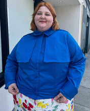 Load image into Gallery viewer, Front view of a size 3X Target x Black History Month cobalt blue wind breaker with large silver snap-closure buttons styled buttoned closed over a white and rainbow floral dress on a size 22/24 model.
