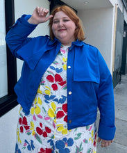 Load image into Gallery viewer, Front view of a size 3X Target x Black History Month cobalt blue wind breaker with large silver snap-closure buttons styled open over a white and rainbow floral dress on a size 22/24 model.
