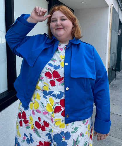 Front view of a size 3X Target x Black History Month cobalt blue wind breaker with large silver snap-closure buttons styled open over a white and rainbow floral dress on a size 22/24 model.