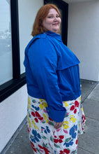 Load image into Gallery viewer, Side view of a size 3X Target x Black History Month cobalt blue wind breaker with large silver snap-closure buttons styled buttoned closed over a white and rainbow floral dress on a size 22/24 model.
