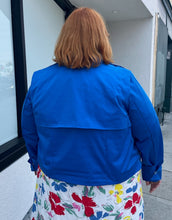 Load image into Gallery viewer, Back view of a size 3X Target x Black History Month cobalt blue wind breaker with large silver snap-closure buttons styled buttoned closed over a white and rainbow floral dress on a size 22/24 model.
