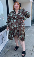 Load image into Gallery viewer, Full-body front view of a size 24 Eloquii brown and light teal mixed geometric floral pattern satin wrap dress with ruffled hem and bishop sleeves styled with black slides on a size 22/24 model. The photo is taken outside in natural lighting.
