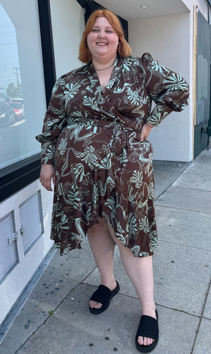 Full-body front view of a size 24 Eloquii brown and light teal mixed geometric floral pattern satin wrap dress with ruffled hem and bishop sleeves styled with black slides on a size 22/24 model. The photo is taken outside in natural lighting.