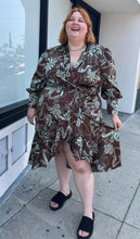 Load image into Gallery viewer, Additional full-body front view of a size 24 Eloquii brown and light teal mixed geometric floral pattern satin wrap dress with ruffled hem and bishop sleeves styled with black slides on a size 22/24 model. The photo is taken outside in natural lighting.
