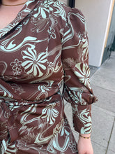 Load image into Gallery viewer, Close up view of the mixed geometric floral pattern of a size 24 Eloquii brown and light teal mixed geometric floral pattern satin wrap dress with ruffled hem and bishop sleeves on a size 22/24 model. The photo is taken outside in natural lighting.
