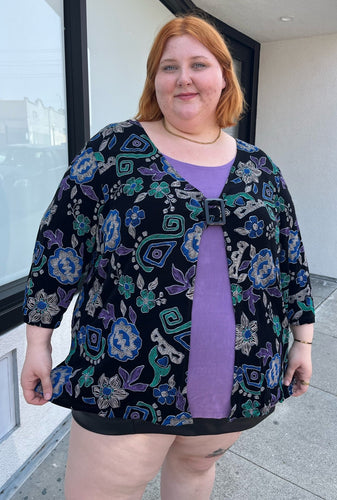 Front view of a size 30/32 Maggie Barnes Vintage black, muted purple, blue, teal, and green mixed pattern floral and geometric blouse with undershirt styled with a pleather mini skirt on a size 22/24 model. The photo is taken outside in natural lighting.