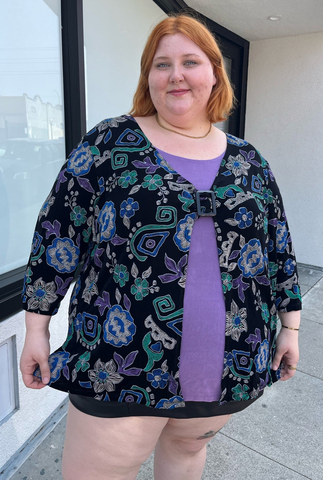 Front view of a size 30/32 Maggie Barnes Vintage black, muted purple, blue, teal, and green mixed pattern floral and geometric blouse with undershirt styled with a pleather mini skirt on a size 22/24 model. The photo is taken outside in natural lighting.