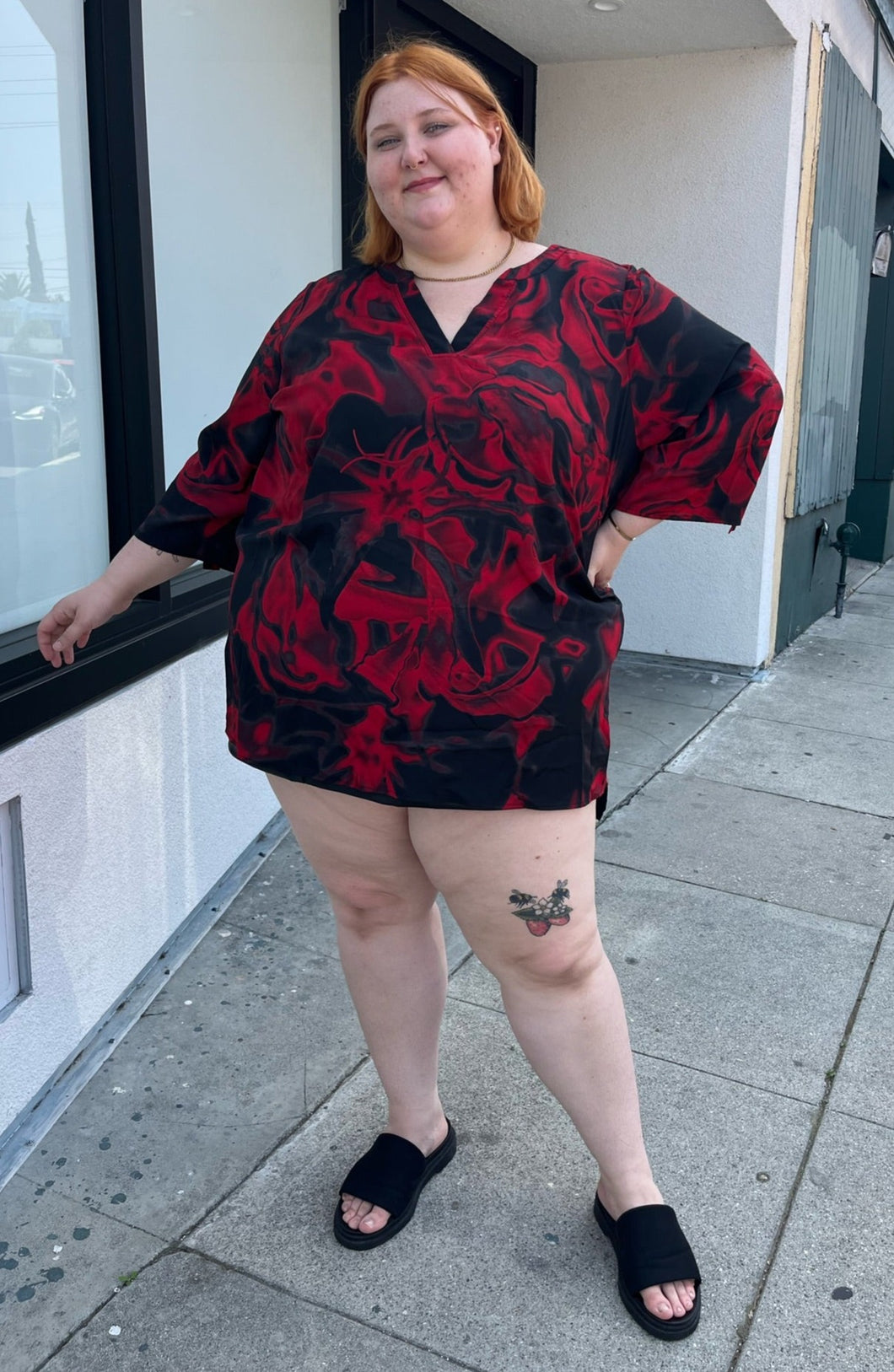 Full-body front view of a size 3X Avenue Vintage red and black psychadelic pattern tunic blouse with a v-neck slit and bell sleeves styled with black slides on a size 22/24 model.