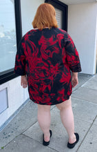 Load image into Gallery viewer, Full-body back view of a size 3X Avenue Vintage red and black psychadelic pattern tunic blouse with a v-neck slit and bell sleeves styled with black slides on a size 22/24 model.
