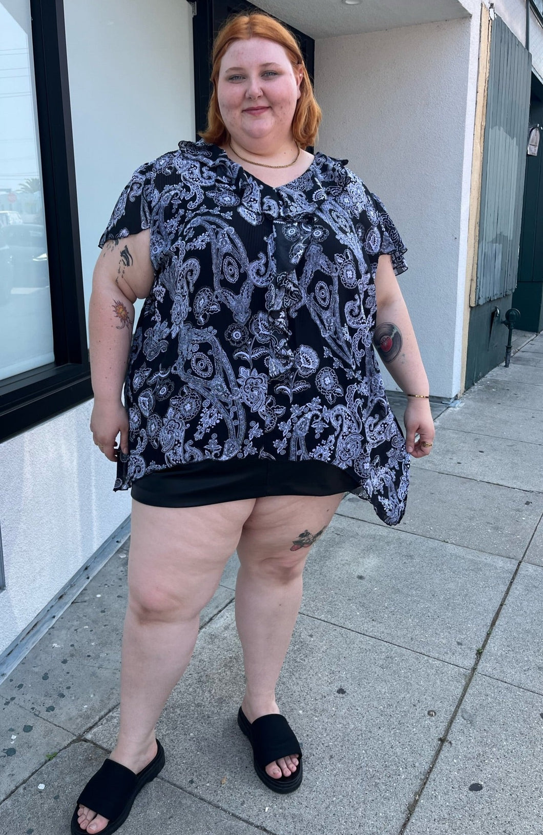 Full-body front view of a size 30/32 Avenue black and white geometric floral flutter sleeve blouse with a ruffle detail at the neckline styled over a black mini skirt with black slides on a size 22/24 model. The photo is taken outside in natural lighting.