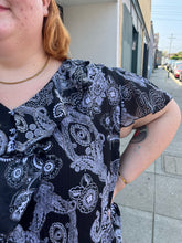 Load image into Gallery viewer, Close up view of the ruffle detail at the neckline and the flutter sleeve of a size 30/32 Avenue black and white geometric floral flutter sleeve blouse with a ruffle detail at the neckline on a size 22/24 model. The photo is taken outside in natural lighting.
