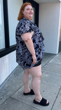 Load image into Gallery viewer, Full-body side view of a size 30/32 Avenue black and white geometric floral flutter sleeve blouse with a ruffle detail at the neckline styled over a black mini skirt with black slides on a size 22/24 model. The photo is taken outside in natural lighting.
