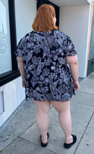 Load image into Gallery viewer, Full-body back view of a size 30/32 Avenue black and white geometric floral flutter sleeve blouse with a ruffle detail at the neckline styled over a black mini skirt with black slides on a size 22/24 model. The photo is taken outside in natural lighting.
