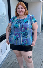 Load image into Gallery viewer, Additional front view of a size 22 Avenue vintage aqua blue, purple, green, and white tropical pattern cap sleeve blouse styled over a black mini skirt on a size 22/24 model. The photo is taken outside in natural lighting.
